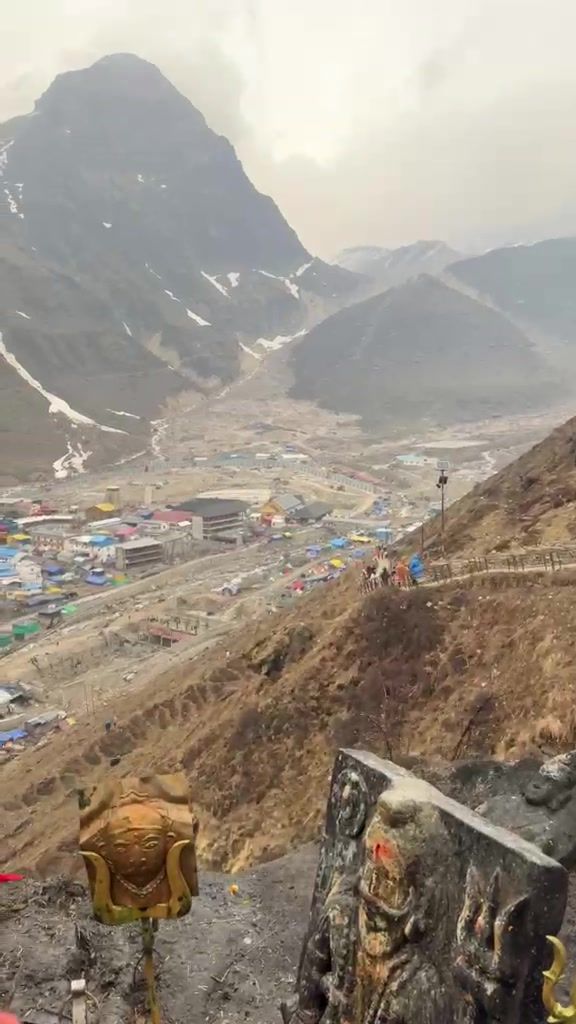 Looking down a brown hillside to the Kedarnath township and valley, with snow-streaked peaks under low cloud.