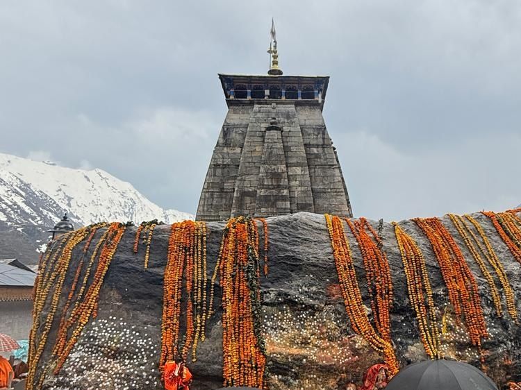 A large boulder draped in marigold garlands, with the stone spire of Kedarnath Temple and snowy peaks behind it.