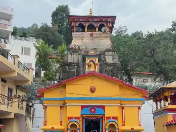A yellow-and-blue temple front below a stone spire, with trees and buildings around it.