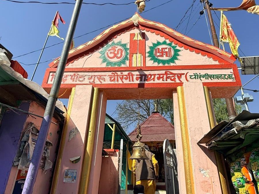 A pink and red temple gateway with a Hindi signboard naming the Guru Chauranginath temple, and a brass bell hanging in the entrance.
