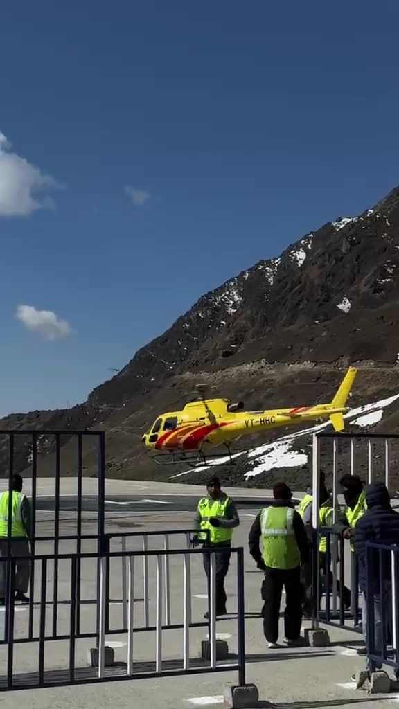 A yellow helicopter on the Kedarnath helipad below a rocky, snow-patched slope, with ground staff behind a barrier.
