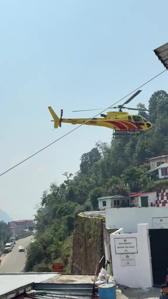 A yellow-and-red helicopter lifting off above the helipad buildings and forested hills.