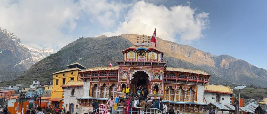 The brightly painted facade of Badrinath Temple among town buildings, with pilgrims at the entrance and mountains under a partly cloudy sky behind.