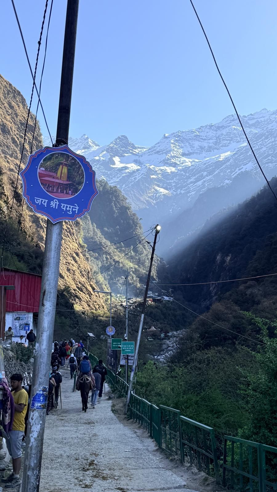 A paved path with walkers climbing into a narrow valley toward snow-capped peaks, beside a round sign reading “Jai Shri Yamune”.