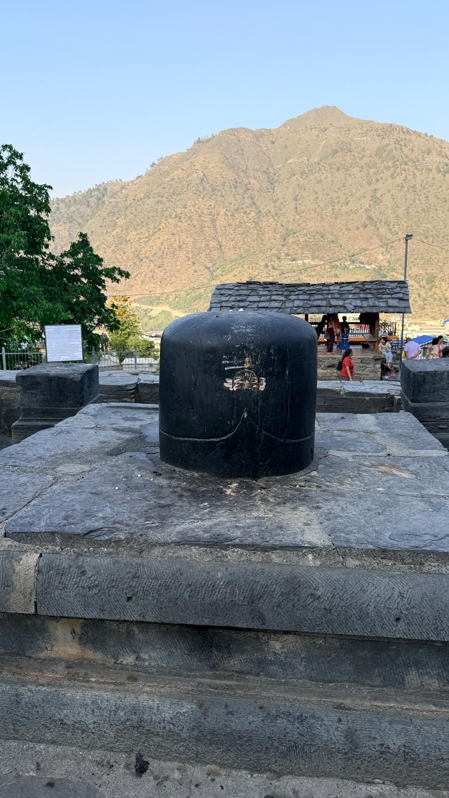 A black stone Shiva linga on a square stone plinth, with a hill and trees behind.