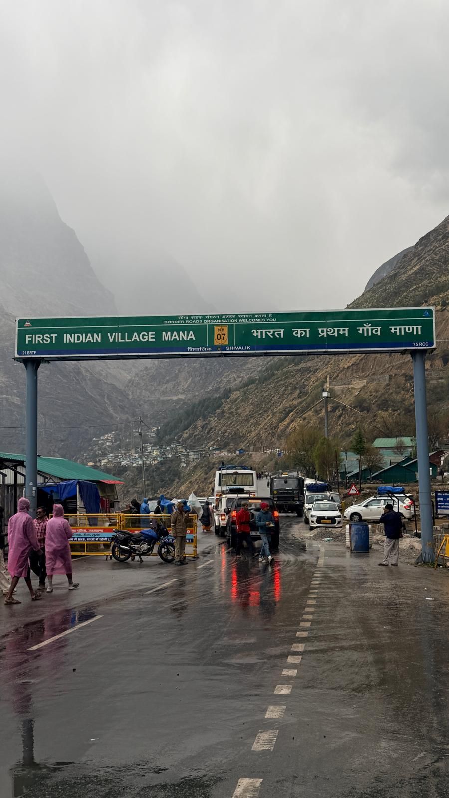 A green roadside gateway reading “First Indian Village Mana” over a wet road, with mist over the mountains.
