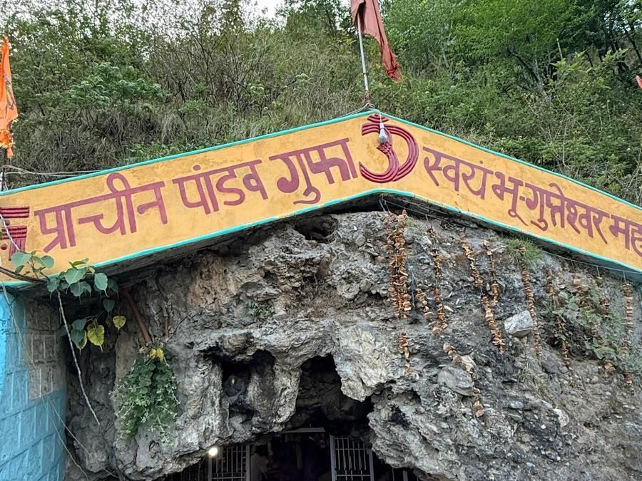 The entrance to a rock cave shrine under a painted Hindi signboard, with a saffron flag above.