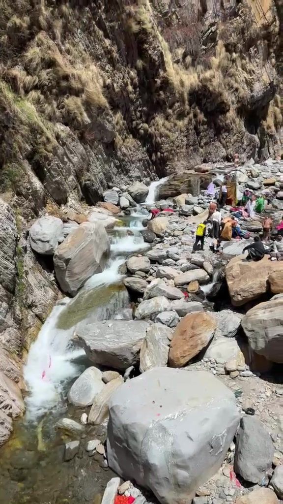 Boulders and a small cascade in a rocky riverbed below steep cliffs, with pilgrims gathered along the banks.
