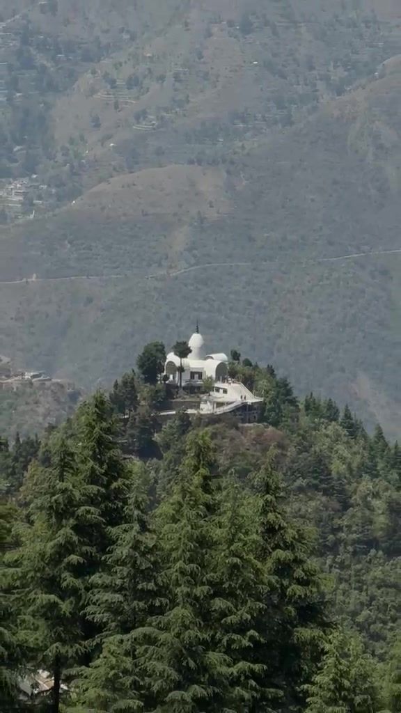A white temple on a hilltop surrounded by pine forest, with hazy ridges behind.
