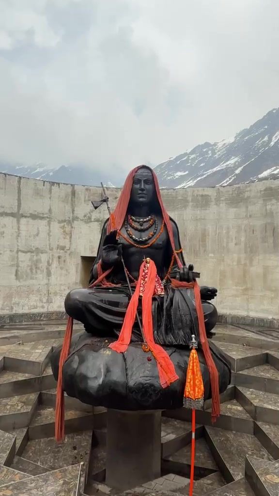 A black stone statue of a seated Adi Shankaracharya, draped in red cloth, in front of a concrete wall and snowy peaks.