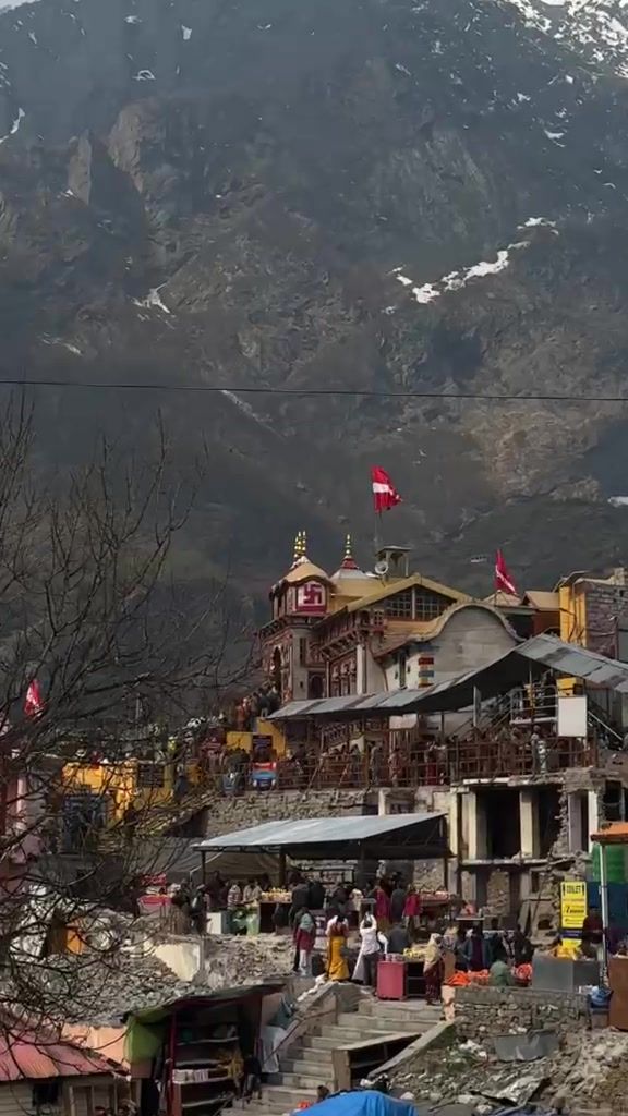 The Yamunotri temple and surrounding buildings on the riverbank below dark, snow-streaked mountains.