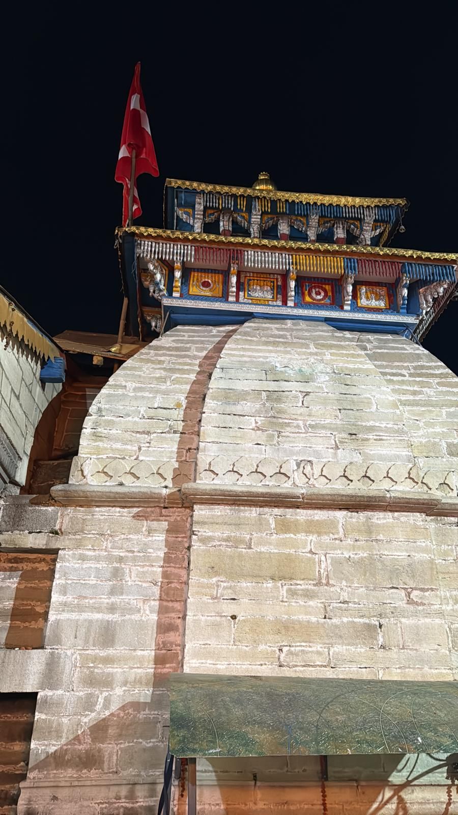 Looking up a pale stone wall to the gilded, painted roof of Badrinath Temple and a flag against the night sky.